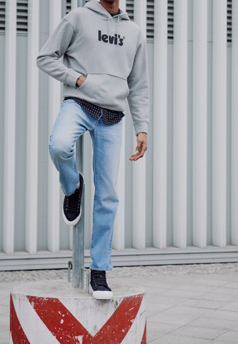 Personne portant un sweat à capuche gris Levi's, un jean bleu clair et des baskets noires, debout avec un pied sur un bloc de béton rayé rouge et blanc.