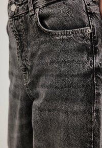 Jeans de mezclilla negra que muestran una textura desgastada, diseño de cinco bolsillos, herrajes metálicos y costuras de contraste a lo largo de las costuras.