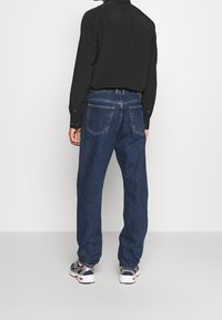Jeans de denim azul oscuro con un ajuste relajado, que presentan dos bolsillos traseros y detalles de costura sutiles, combinados con una camisa negra de manga larga.
