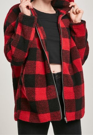 Osoba ubrana w czerwoną i czarną polarową kurtkę w kratę z zamkiem, trzymająca kołnierz, a pod spodem czarny crop top i spodnie.