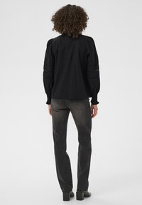 Personne avec des cheveux bouclés portant un haut noir à manches longues et un jean noir décoloré, debout de dos sur un fond clair.