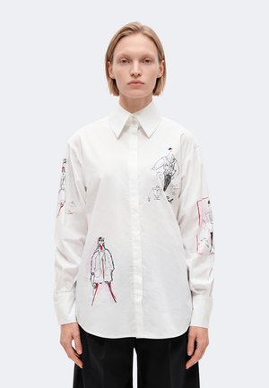 Un mannequin se tient devant un fond clair, portant une chemise blanche ornée de croquis de mode et de touches rouges, associée à un pantalon noir.