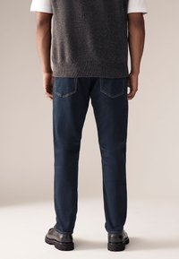 Tummansininen farkkuhousut, joissa suorasyöttö, kaksi takataskua ja kontrastiväriset saumat, yhdistettynä harmaaseen neulottuun liiviin. Mustat saappaat näkyvissä.