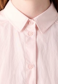 Gros plan d'une chemise rose clair à col boutonnée jusqu'au cou, montrant trois boutons ainsi qu'une partie de l'encolure et du torse.