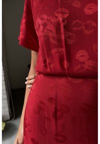 Robe rouge avec un motif discret d'empreintes de lèvres, manches courtes et une main posée sur la zone de la taille au niveau des hanches.