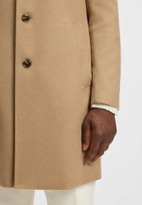 Cappotto di lana beige con tre bottoni, superficie testurizzata e tasche laterali. Un polsino in maglione color crema è visibile spuntare dalla manica.