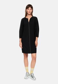 Schwarzes Kleid zum Zuziehen mit V-Ausschnitt, gerafften Ballonärmeln und kontrastierenden Nähten, kombiniert mit silbernen Sneakers und gelb gestreiften Socken.