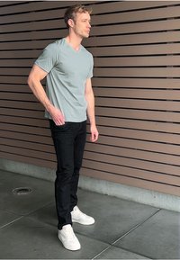 Muž stojící s rukou v kapse, oblečený v světle šedém tričku, černých džínách a bílých teniskách, před hnědou horizontální laťkovou stěnou.