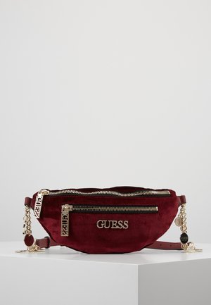 Sac banane Guess en velours rouge foncé avec zips dorés, logo et détails de chaîne, posé sur une surface blanche.