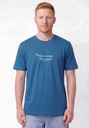Man with short red hair wearing blue T-shirt with white text in German "Was muss, das muss" standing against plain white background.