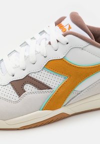 White sneaker with yellow suede accents, brown details, and perforated design. Features white laces and a light brown cushioned collar.