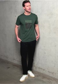 Zelené bavlněné tričko s grafickým potiskem, spárované s černými kalhotami a bílými teniskami. Jednoduchý design, volný střih a hladká textura.