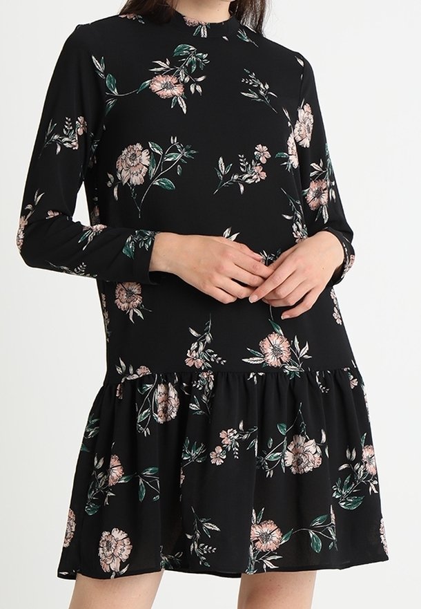 Femme portant une robe noire à manches longues avec un motif floral rose et vert, dotée d'un col montant et d'un ourlet froncé à la jupe.