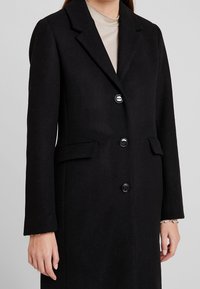 Schwarzer Wollmantel mit Reverskragen, einreihigen Knöpfen, zwei seitlichen Taschen und langen Ärmeln mit dezentem Manschettendetail.