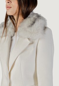 Manteau en mélange de laine blanc avec un col en fausse fourrure gris clair et pelucheux. Présente un revers pointu et une silhouette structurée.