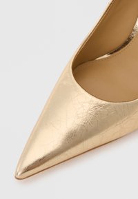 Pointed toe gold metallic leather high heel shoe shown in close-up on white background.