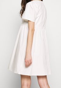 Ljusbeige kortärmad klänning med figursydd överdel och utsvängd kjol, tillverkad av bomull. Har rund halsringning och sidofickor.