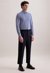Uomo in piedi che indossa una camicia a righe blu e bianche con bottoni, pantaloni scuri navy, calze nere e scarpe eleganti nere su uno sfondo semplice.