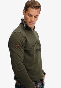 Oliwkowy sweatshirt z wypukłym napisem "Superdry Athletics" z przodu, z teksturowanym wykończeniem i kołnierzykiem z koszuli w kratę pod spodem.