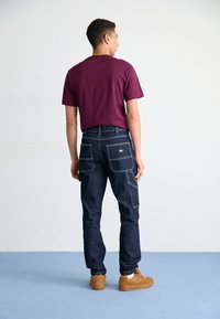 Dark blue denim pants with a relaxed fit, featuring multiple pockets and contrasting stitching. Paired with a maroon short-sleeve shirt.