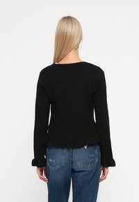 Eine Frau mit langen blonden Haaren trägt einen schwarzen Pullover mit langen Ärmeln und Rüschekanten sowie blaue Jeans und steht mit dem Rücken zur Kamera.
