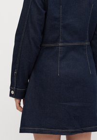 Πίσω όψη ενός ατόμου που φοράει ένα σκούρο μπλε τζιν φόρεμα με ορατές ραφές και κουμπωτές μανσέτες στα μακριά μανίκια.