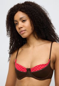 Καφέ σουτιέν με κόκκινα διακοσμημένα πάνω μέρη φλυτζανιών. Διαθέτει ρυθμιζόμενες τιράντες και λεπτομέρεια με φιόγκο στο κέντρο. Ομαλή υφή με αντιθετικά χρώματα.