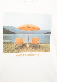 Valkoinen t-paita, jossa on grafiikka kahdesta oranssista loungetuolista ja samanvärisestä aurinkovarjosta järven äärellä vuorten taustalla.