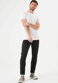 Camiseta blanca de algodón combinada con unos vaqueros negros y zapatillas altas grises. El modelo está en una pose relajada contra un fondo simple.