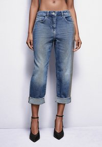 Jeans de mezclilla azul con un corte relajado, dobladillos enrollados, bolsillos delanteros y traseros, y un leve desgastado. Llevados con zapatos de tacón negro.