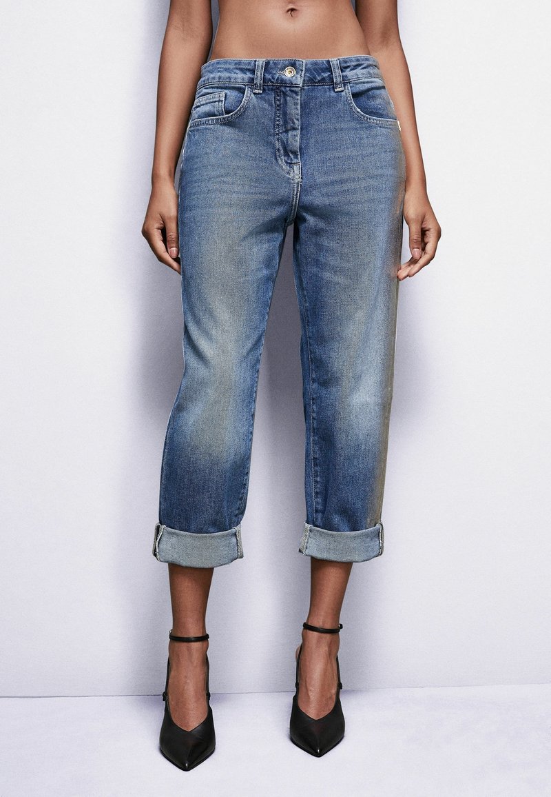 Jeans de mezclilla azul con un corte relajado, dobladillos enrollados, bolsillos delanteros y traseros, y un leve desgastado. Llevados con zapatos de tacón negro.