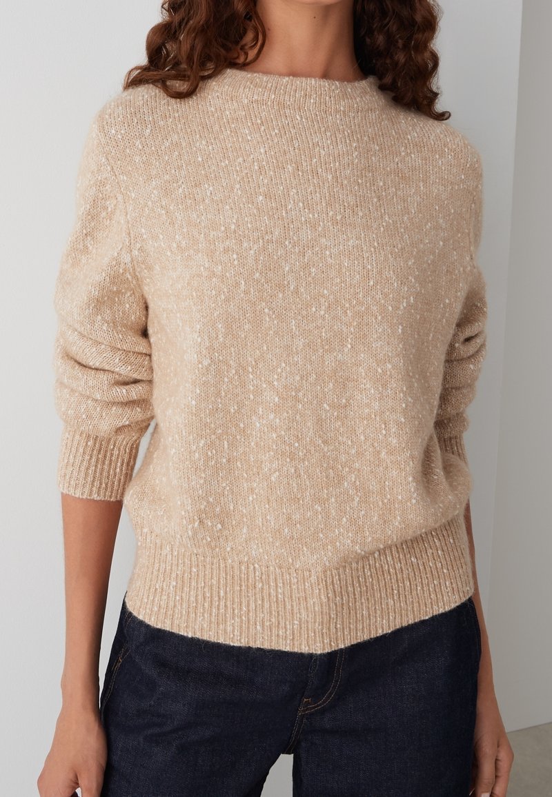 Pull en maille beige mouchetée avec des poignets et un ourlet côtelés, porté avec un jean bleu foncé, visible de la base du cou jusqu'aux hanches.