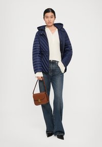 Žena nosi tamnoplavi patchwork kaput, bijeli rebrasti sv sweater, plave traperice, crne cipele na špic, dok drži smeđu kožnu torbu.