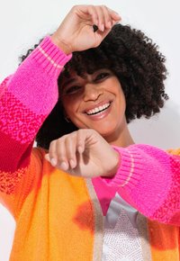 Persona sorridente con capelli ricci che indossa un maglione colorato arancione e rosa, sollevando le braccia in modo giocoso contro uno sfondo bianco uniforme.