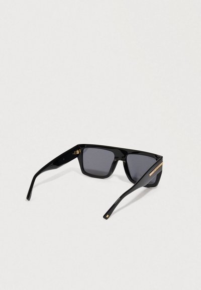 Gafas de sol negras de forma rectangular con lentes oscuros, patillas gruesas y un pequeño acento dorado en la patilla, contra un fondo claro.