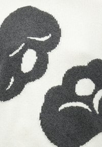 Pleteni pulover z belo podlago in velikimi temnosivimi risankastimi obrazi, zaobljenimi oblikami z izrazitimi očmi in poudarki na ustih.