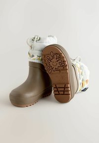 Bruine rubberen laarzen met een witte canvas bovenkant, een patroon rondom en een trekkoordsluiting. Geribbelde zool met een glimlach-splash detail.