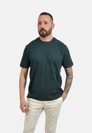 Uomo con capelli corti e scuri e barba, che indossa una camicia verde scuro a maniche corte e pantaloni chiari, con tatuaggi sul braccio destro.