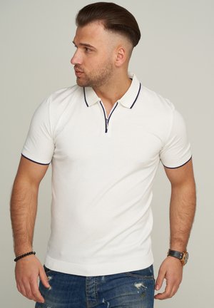 Weißes Poloshirt mit kurzem Arm und Reißverschlusskragen, akzentuiert mit marineblauen Details am Kragen und an den Ärmeln, kombiniert mit einer abgetragenen blauen Jeans.
