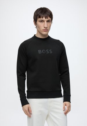 Homme debout devant un fond uni, portant un sweat noir avec le texte "BOSS" sur la poitrine et un pantalon blanc, face à l'objectif avec une expression neutre.
