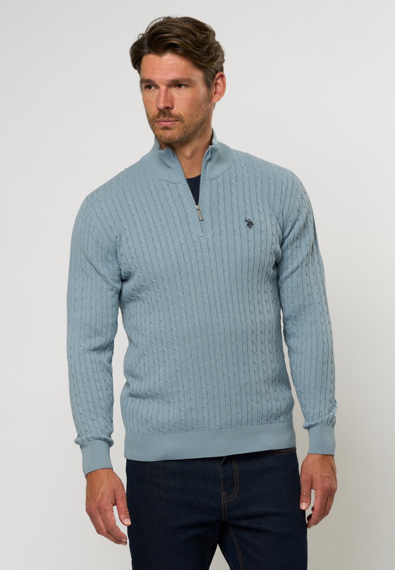 Gaiši zils piedurkņu džemperis ar virsrakstu, kas izgatavots no kabeles adījuma. Ietver augstu apkakli un ribota piedurknes, apvienojoties ar tumšām džinsām. Nav redzamu logo.