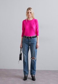 Pielāgots rozā ilgajai piedurknēm tops, zili nolietoti džinsi ar plīvējiem, melna josta un melni ankletu zābaki. Modelis tur mazu melnu rokassomu.