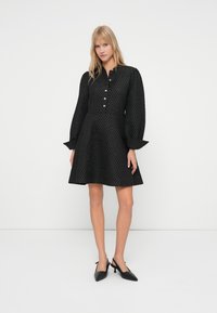 Sieviete stāv, ģērbusies melnā struktūrētā garajās piedurknēs kleitā ar pogām un melnām šaurām slingback kurpēm uz vienkrāsaina baltā fona.