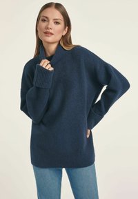 Granatowy, oversize sweter dzianinowy z wysokim kołnierzem, prążkowanymi mankietami i teksturowanym wykończeniem, w zestawieniu z jasnoniebieskimi dżinsami.