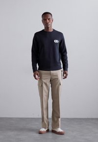 Fekete pulóver mellkasi logóval, bézs cargo nadrággal, oldalzsebekkel, és fehér edzőcipőkkel, barna talppal.