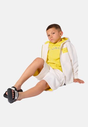 Garçon assis par terre portant un sweat à capuche blanc avec doublure jaune, un t-shirt jaune, un short blanc et des sandales à glissière noires avec des brides blanches.