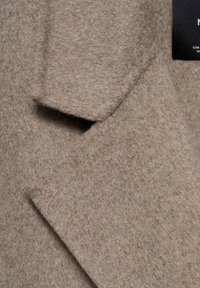 Beige villakangas takki pehmeällä, teksturoidulla pinnalla. Ominaisuuksiin kuuluu lovattu kaulus ja yksi tasku, joissa on siistit linjat räätälöidyn ilmeen saavuttamiseksi.