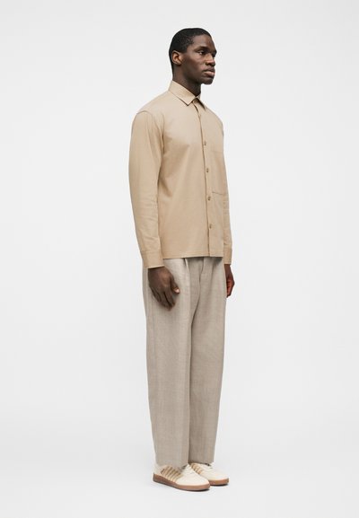 Άνδρας όρθιος, φορώντας μπεζ πουκάμισο με κουμπιά, ανοιχτόγριζο παντελόνι με φαρδιά πόδια και λευκά αθλητικά παπούτσια με καφέ σόλες, μπροστά σε απλό λευκό φόντο.