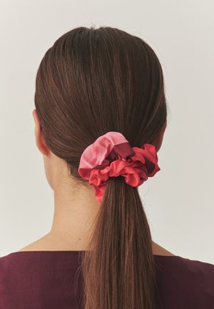 Zijden scrunchie in verschillende tinten roze, met een gerimpeld ontwerp. Het haar is naar achteren gebonden in een strakke paardenstaart, waarbij het accessoire wordt benadrukt.
