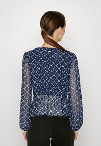 Blusa blu navy con maniche lunghe trasparenti e vita a peplum. Presenta un motivo geometricamente blu chiaro su tutta la superficie e un colletto rotondo.